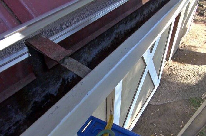 A close-up view of a house’s roof edge shows a dirty, clogged gutter filled with dark debris and leaves above a window—highlighting the need for Gutterglove and its RiserStep Technology.
