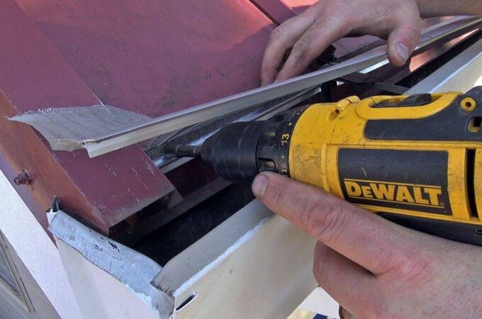 A person uses a yellow DeWalt power drill to secure stainless steel micro-mesh with a metal strip to the edge of a red metal roof. Their hands are visible holding the drill and materials, ideal for DIYers shopping at Home Depot with same-day pickup.