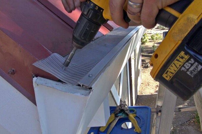 A person uses a DEWALT power drill to attach a Gutter Guard by Gutterglove with RiserStep Technology to the edge of a roof, with tools visible nearby on a blue platform.