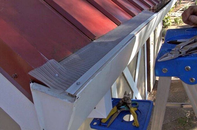 A person is installing or cleaning a Gutter Guard by Gutterglove on a house with a red metal roof. Several tools, including pliers and cutters, are on a blue step ladder next to the gutter.