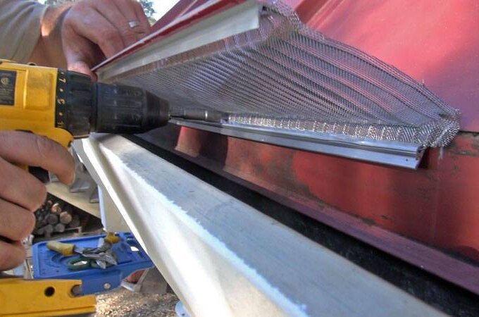 A person uses a power drill to attach a metal mesh guard with RiserStep Technology to the edge of a roof above a gutter, likely for gutter protection or pest prevention.