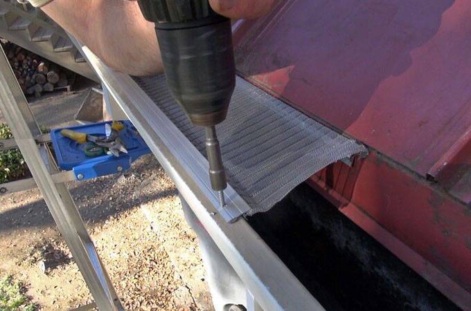 Close-up of a person using a power drill to attach the Gutter Guard by Gutterglove onto the edge of a roof, with a ladder and tools nearby.