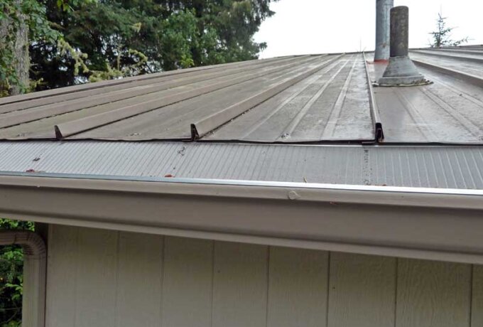 A close-up view of a house roof with a Gutter Guard by Gutterglove installed, preventing debris from entering the gutter; trees are visible in the background. Same-day pickup is available.
