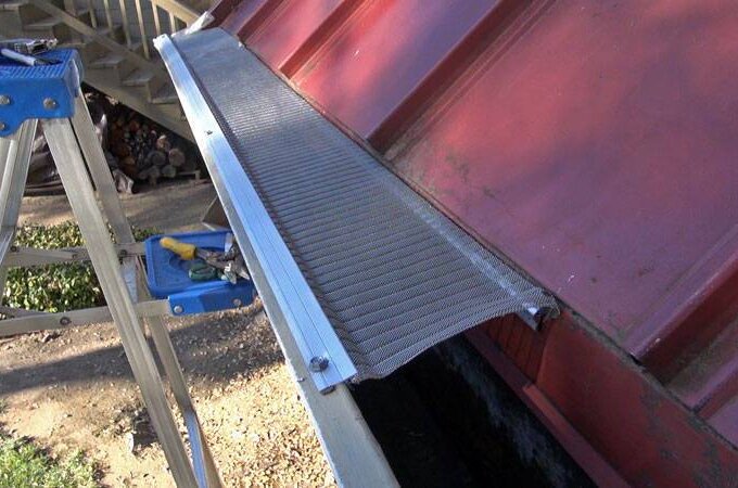 A stainless steel micro-mesh gutter guard is partially installed along the edge of a red metal roof. A blue stepladder with tools sits nearby, with trees and firewood visible in the background.