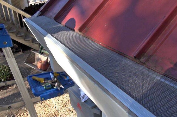 Close-up of a metal roof with Gutterglove gutter guard featuring RiserStep Technology installed along the edge. A blue ladder with tools rests beside the roof, and fallen leaves are scattered on the ground below.