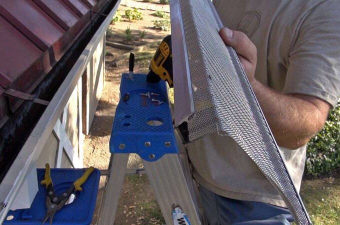 A person stands on a ladder installing Gutter Guard by Gutterglove on the edge of a house roof, holding a tool and a drill, with pliers visible on the ladder platform. Available for same-day pickup at Home Depot.
