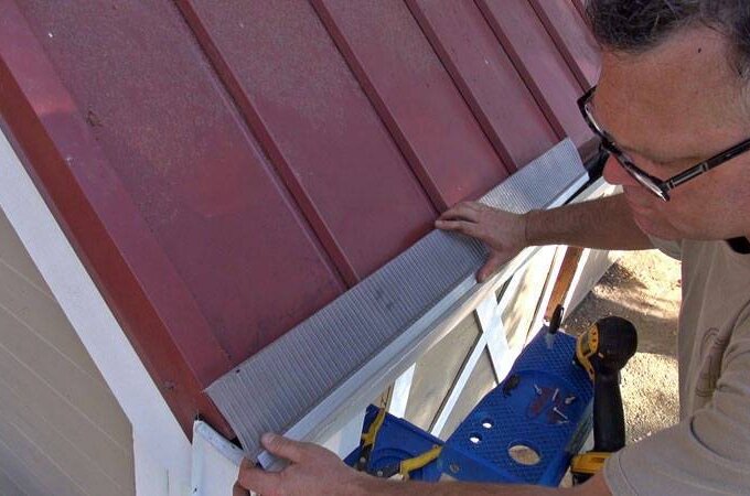 A person wearing glasses uses a tool to secure metal flashing along the edge of a red metal roof above a white-framed window, showcasing Gutterglove installation with materials available for same-day pickup at Home Depot.