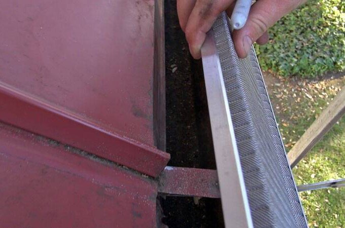 A person stands on a ladder installing a Gutterglove metal gutter guard along the edge of a red roof, with a garden visible below—perfect for easy home upgrades from Home Depot with same-day pickup.