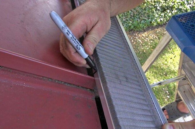 A person uses a black Sharpie marker to mark a piece of Gutterglove stainless steel micro-mesh gutter guard resting on the edge of a red metal roof, with a blue ladder and green grass visible nearby.