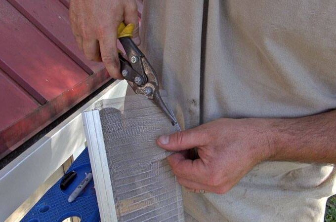 A person uses tin snips to cut a piece of mesh screen with RiserStep Technology, likely for a Gutter Guard by Gutterglove, while standing next to a house roof.
