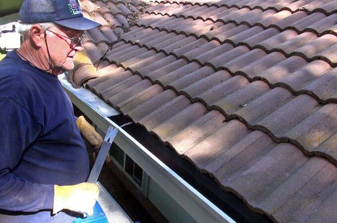 An older man wearing gloves and a cap stands on a ladder next to a tiled roof, cleaning debris from the house’s rain gutter with a tool. Gutterglove products are available at Home Depot for same-day pickup.