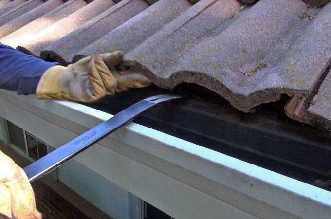 A person wearing gloves uses a pry bar to lift a curved roof tile above a house gutter, possibly installing Gutter Guard by Gutterglove with stainless steel micro-mesh for effective roof or gutter maintenance.