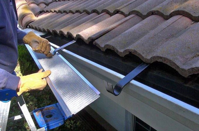 A person wearing gloves installs a Gutter Guard by Gutterglove over a house gutter, using a tool to secure it under the edge of the roof’s tiles, similar to products found at Home Depot.