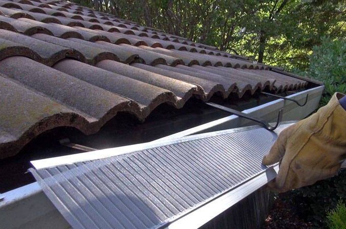 A person wearing gloves installs a stainless steel micro-mesh guard over a house’s rain gutter, which runs along the edge of a tiled roof with trees visible in the background.