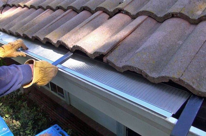 A person wearing gloves installs a Gutter Guard by Gutterglove beneath the edge of a tiled roof, using tools to secure it in place above a white gutter.