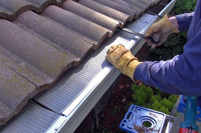 A person wearing gloves installs a metal gutter guard with RiserStep Technology along the edge of a tiled roof, using tools placed nearby on a step ladder. Green plants are visible below, showcasing a project inspired by Home Depot solutions.