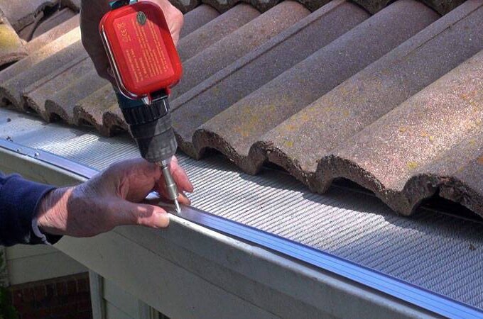 A person uses a power drill to install a metal gutter guard with RiserStep Technology along the edge of a tiled roof, securing it in place to prevent debris from entering the gutter.