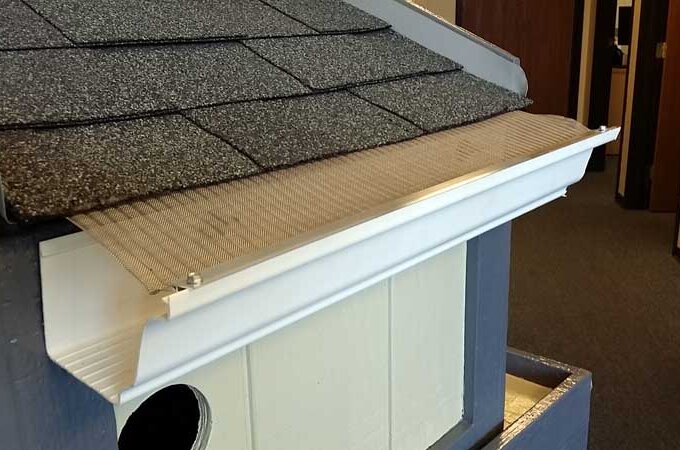 A close-up of a roof section with asphalt shingles and a white gutter guard featuring stainless steel micro-mesh, showing how it covers the gutter to prevent debris from entering.