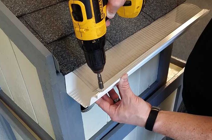 A person uses a yellow power drill to attach a white piece of trim featuring RiserStep Technology to the edge of a small roofed structure with gray shingles and blue-gray trim.