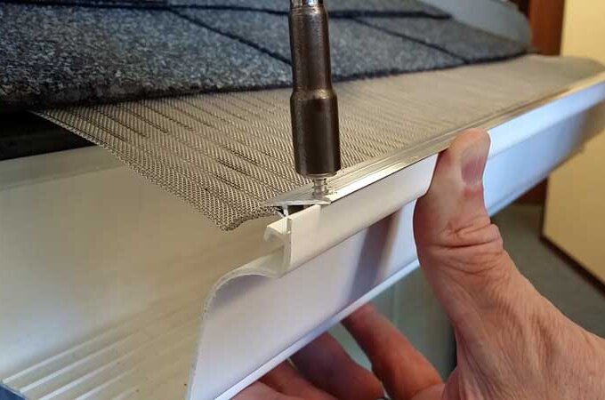 A person installs a stainless steel micro-mesh gutter guard with RiserStep Technology on a roof, using a power tool to secure it onto the edge of a white rain gutter beneath gray shingles.
