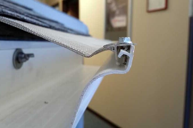 Close-up of the edge of a white retractable awning with a metal clamp securing the fabric to the frame, taken indoors with a blurred background—available for same-day pickup at Home Depot.