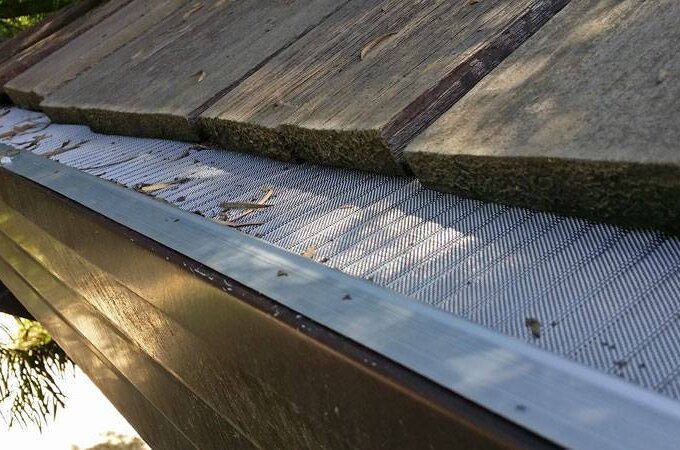 Close-up of a house roof with a Gutterglove metal gutter guard featuring RiserStep Technology, preventing leaves and debris from entering the gutter. Some small twigs and leaves rest on top of the mesh screen near the roof’s edge.