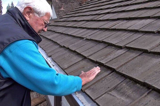An older man in a blue jacket inspects and touches the shingles on a sloped roof, possibly checking for damage or wear, while considering stainless steel micro-mesh for added protection.