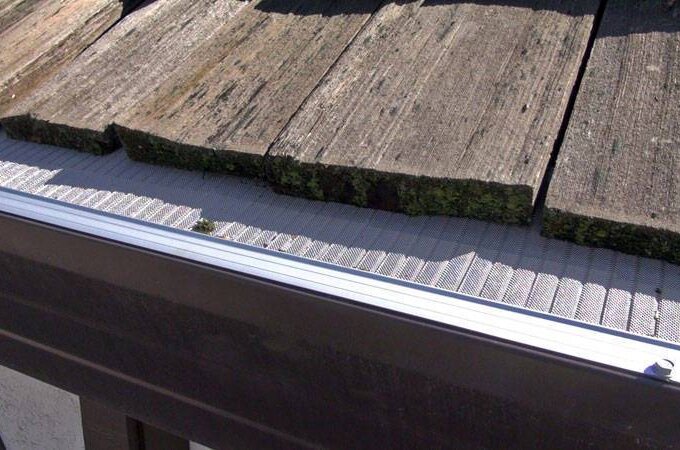 Close-up of a roof with weathered wooden shingles, some with moss, above a metal gutter fitted with a Gutterglove stainless steel micro-mesh guard to prevent debris from entering.