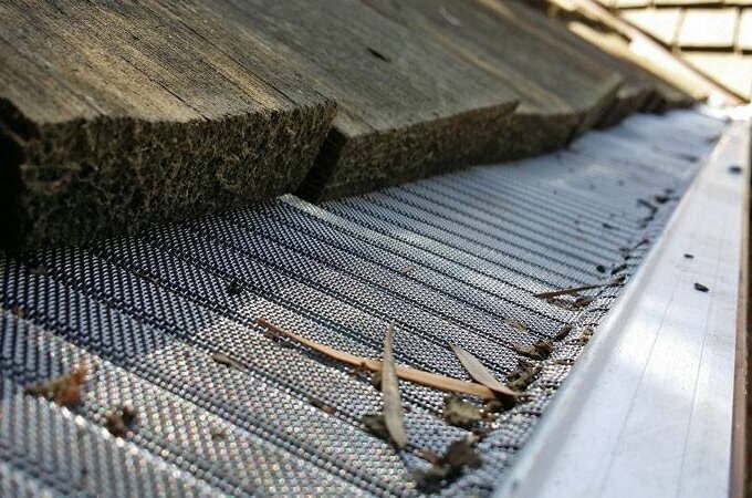 Close-up of a roof edge with wooden shingles and a metal mesh Gutter Guard by Gutterglove, with small debris and leaves caught on top. Same-day pickup available.