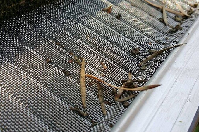 Close-up of a metal gutter guard with small debris, including leaves and dirt, resting on its mesh surface. The gutter guard featuring RiserStep Technology is installed along the edge of a roof.