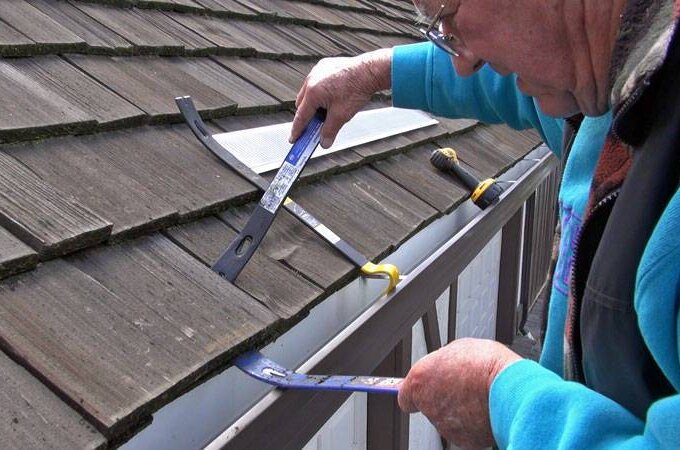 A person in a blue jacket uses tools to repair or install shingles on a sloped roof near the edge above a white gutter, where Gutter Guard by Gutterglove provides added protection.