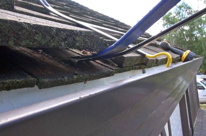 Close-up of roof tiles being lifted with tools, possibly for repairs or installation, next to a gutter fitted with stainless steel micro-mesh on a sloped roof. Trees and cars are visible in the background; same-day pickup available.