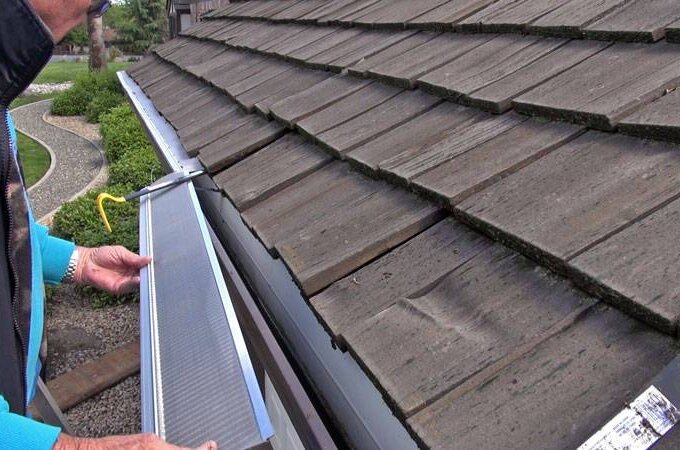 A person installs a stainless steel micro-mesh gutter guard along the edge of a roof covered with wooden shingles, helping to keep debris out of the gutter. Shrubs and a gravel path are visible in the background.