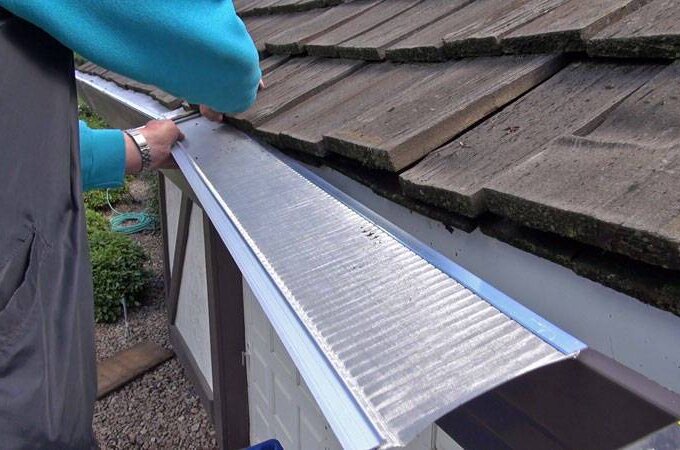 A person installs a Gutter Guard by Gutterglove with RiserStep Technology along the edge of a shingled roof using a screwdriver. Only the hands and arms are visible, with the house's exterior and yard below.
