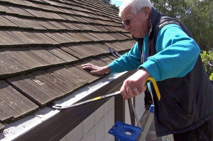 An older man wearing sunglasses and a blue jacket repairs the edge of a wooden shingle roof, using a crowbar near the white garage door. Trees are visible in the background, and stainless steel micro-mesh is installed along the roofline.