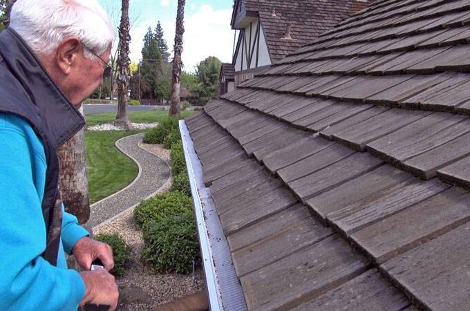 An elderly man in a blue jacket inspects the rain gutter along the edge of a shingle roof, with a landscaped yard and curving stone path behind him, considering adding Gutter Guard by Gutterglove’s stainless steel micro-mesh for protection.